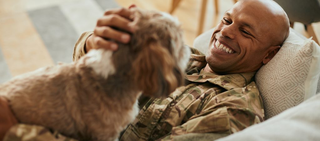 Smiling service member in camouflage uniform relaxing on a couch while petting a fluffy brown and white dog.